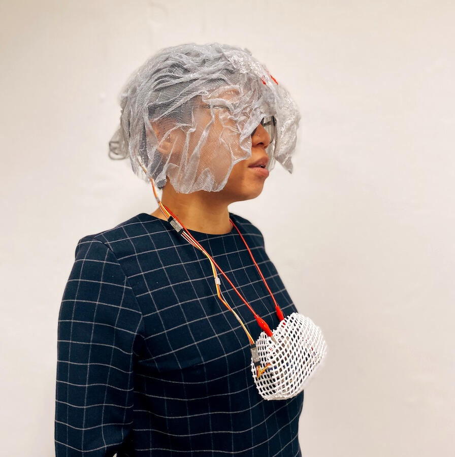 Sam in a navy checkered top, with mesh covering their head and face; this is connected to electronics encased in a white netting pouch hung over their neck